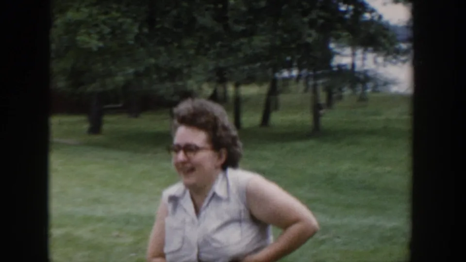 A smiling woman with glasses stands in a green Wisconsin yard in 1953, wearing a sleeveless blouse against a tree-filled background.