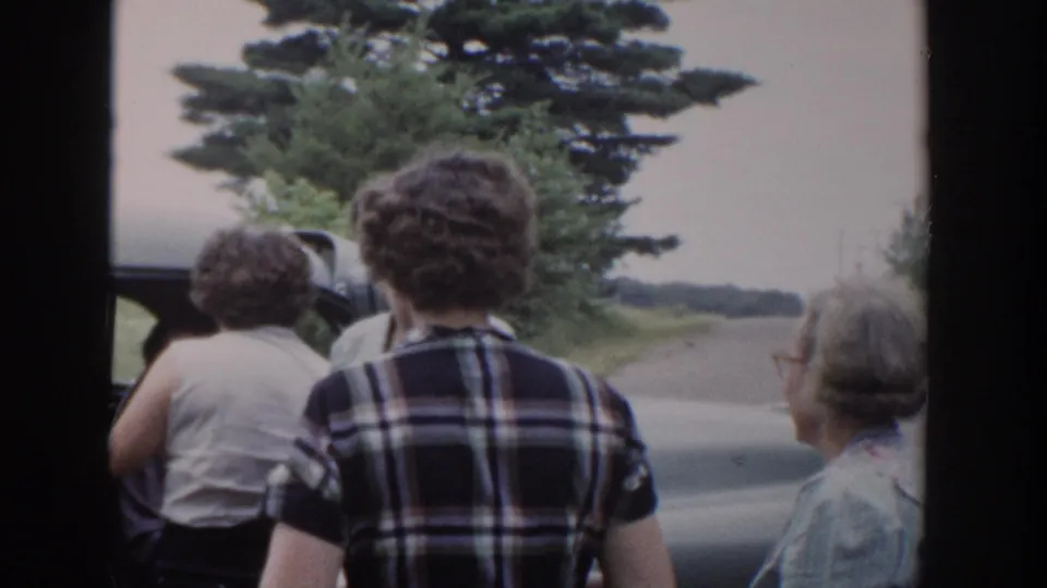 Women in 1950s attire stand close to an open car door along a quiet Wisconsin roadside.