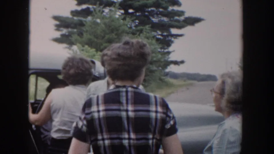 Handheld 8mm frame of women gathered near a car, with a rural Wisconsin road and trees in the background.