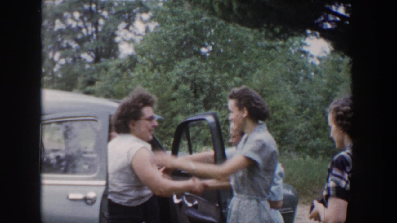 In 1953 Wisconsin home-movie footage, women greet beside a light-colored car with an open door and leafy trees behind them.