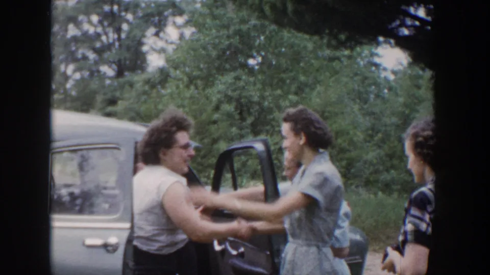 In 1953 Wisconsin home-movie footage, women greet beside a light-colored car with an open door and leafy trees behind them.