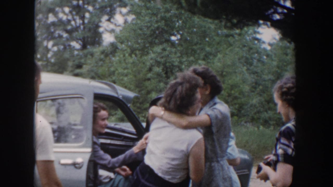 A woman in a white sleeveless top hugs a woman in a blue dress beside a car in supported 1953 Wisconsin home-movie footage.
