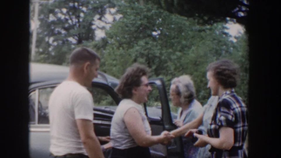 A small group greets one another by the open car door, with a man in a white T-shirt and women in period dresses visible.
