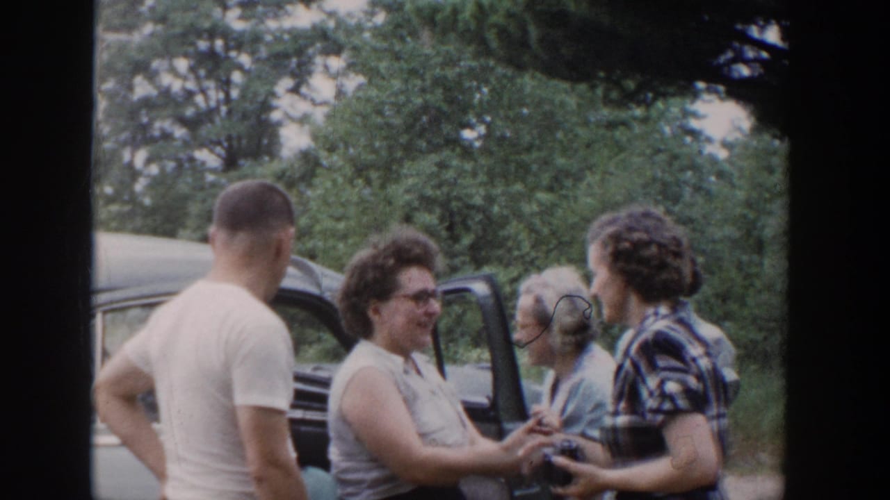 1953 Wisconsin home-movie frame showing adults gathered by a blue car, with women smiling and a man turned away near the open door.