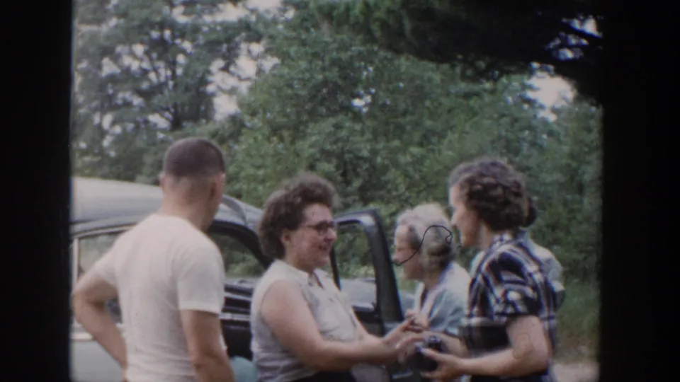 1953 Wisconsin home-movie frame showing adults gathered by a blue car, with women smiling and a man turned away near the open door.