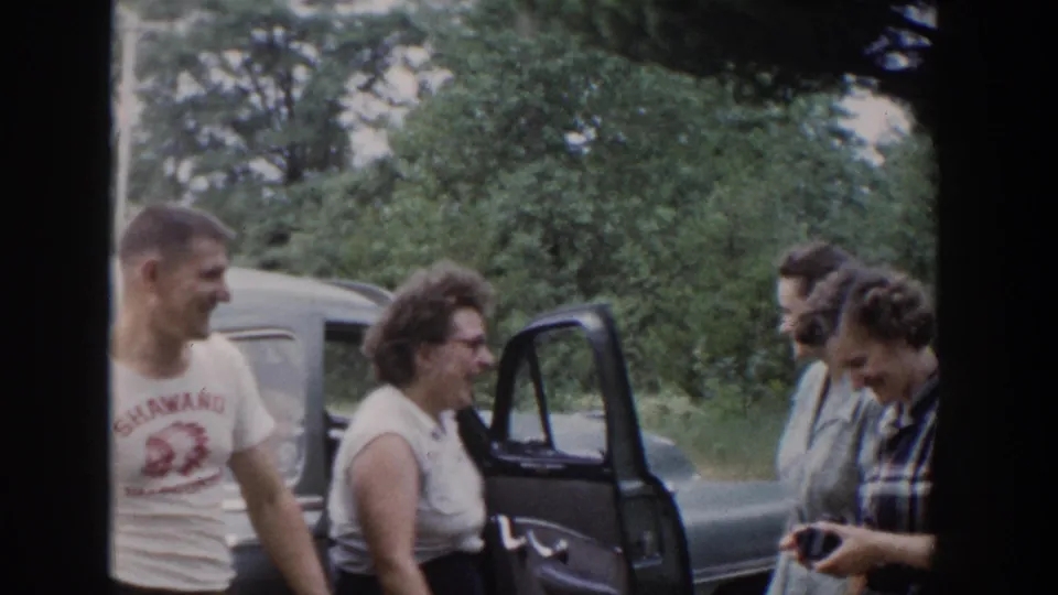 Adults in 1953 Wisconsin appear to laugh and chat near a blue car, with a small camera or object visible in one woman's hands.
