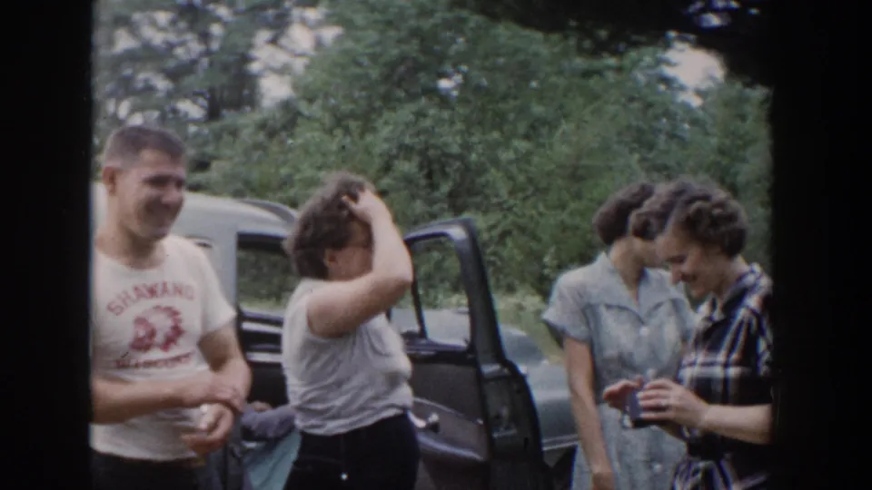 1953 Wisconsin home-movie frame showing women examining a small object near a blue car while a man in a printed T-shirt stands nearby.
