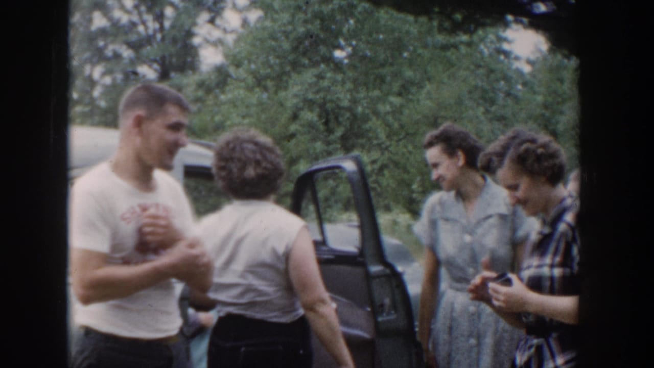 A man smiles while women talk near an open car door and trees.
