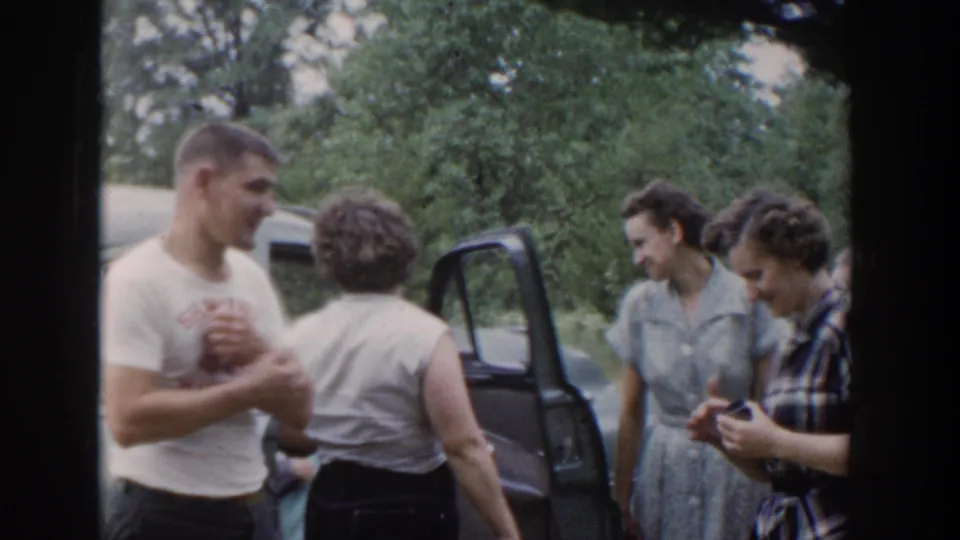 A smiling man in a printed shirt stands with women near a classic car in wooded 1953 Wisconsin footage.