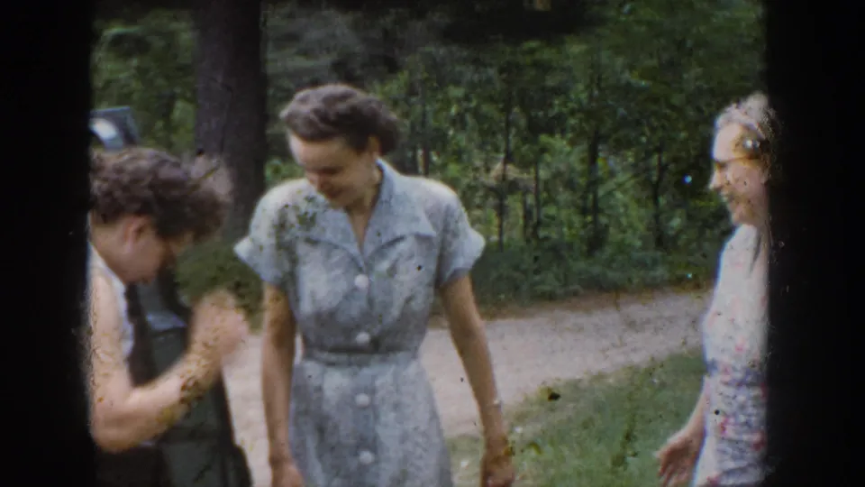 Women in 1950s dresses laugh and gesture near the parked car during the Wisconsin outdoor gathering.