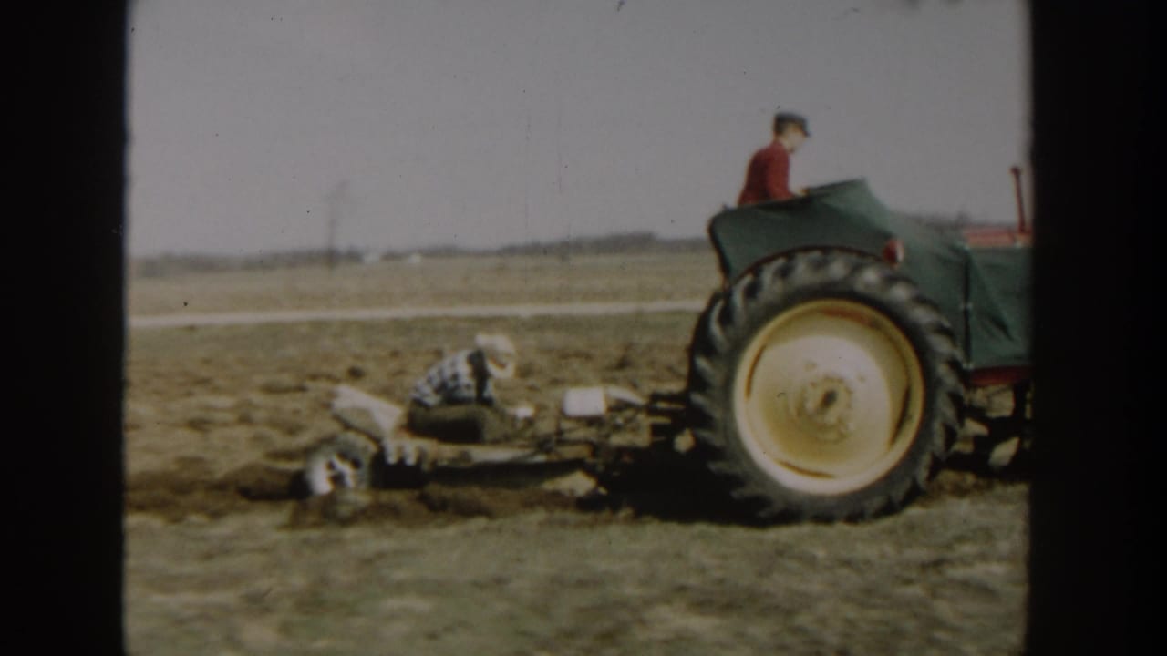 1954 Wisconsin Dells home-movie frame showing a green tractor near a crouching man working beside plow equipment in open farmland.