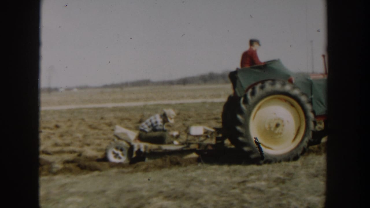 Tractor wheel and driver pass close to a crouching worker and plow attachment.