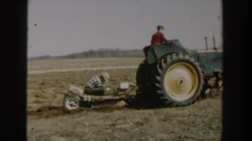 1954 Wisconsin Dells footage shows a man driving a green tractor while another man crouches near field equipment on open farmland.