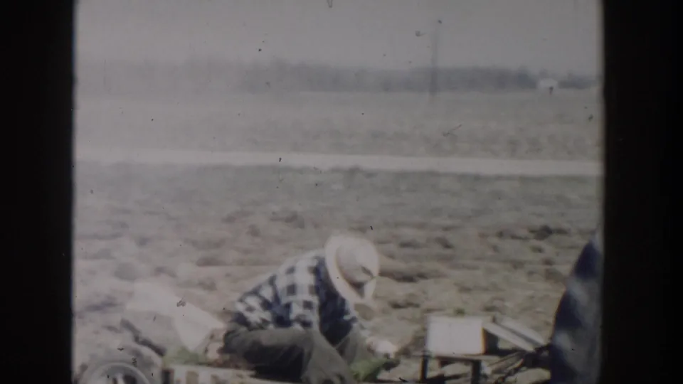 A 1954 Wisconsin Dells home-movie frame centers on a crouching worker in a plaid shirt and straw hat beside farm equipment in an open field.