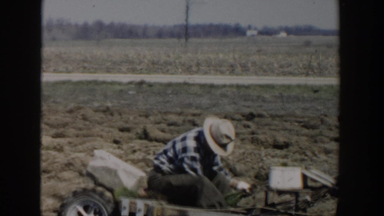 In Wisconsin Dells in 1954, a man in a straw hat and plaid shirt crouches beside small farm equipment in an open field.