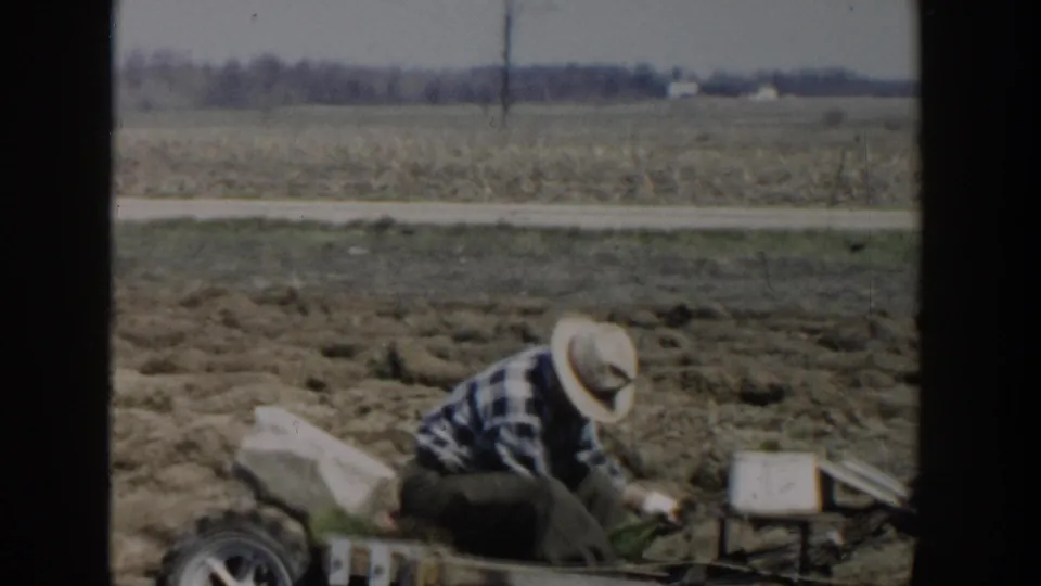 In Wisconsin Dells in 1954, a man in a straw hat and plaid shirt crouches beside small farm equipment in an open field.