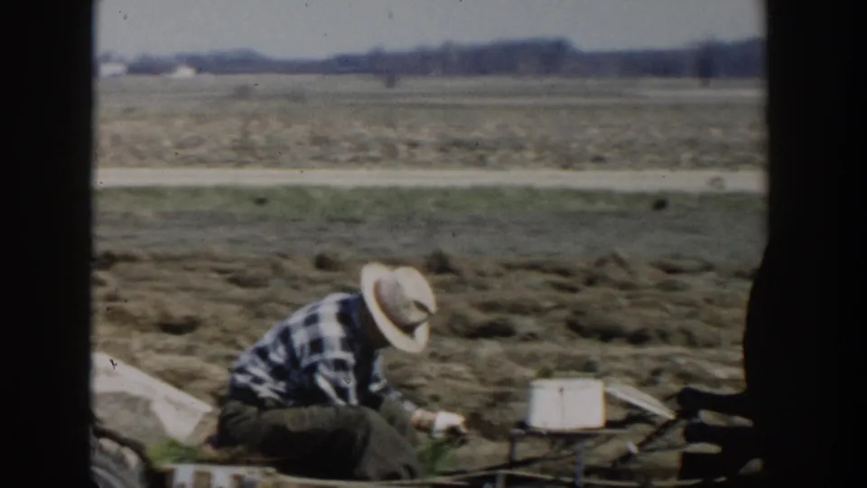 A 1954 Wisconsin Dells farm scene shows a man crouched near a small implement, with fields and a road behind him.