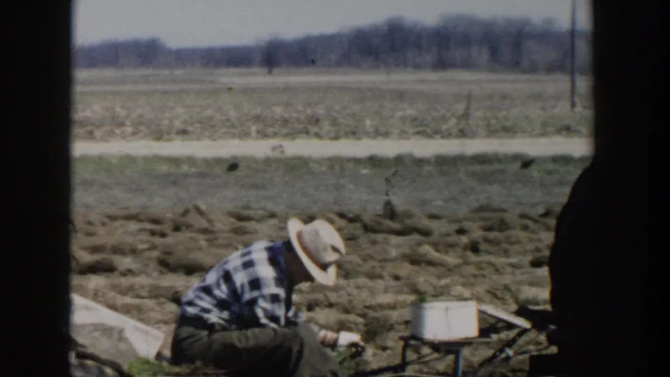In the 1954 Wisconsin Dells home movie, the man works close to the soil beside equipment as fields and trees fill the background.