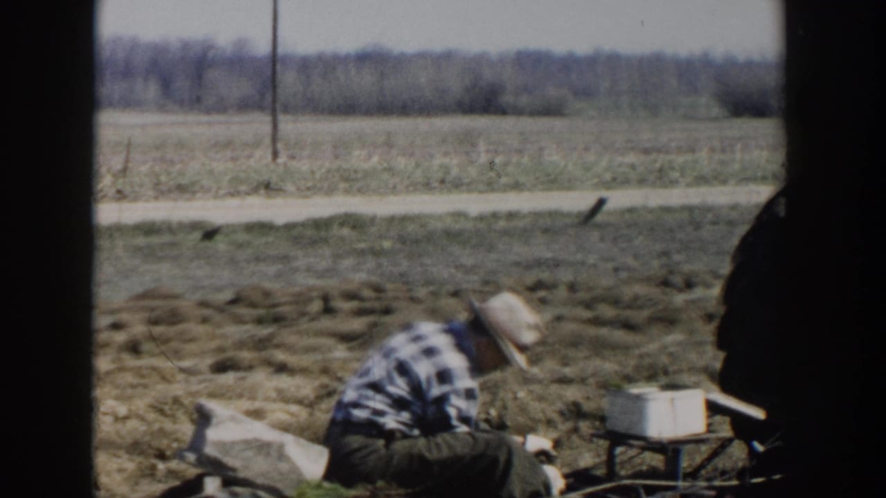 Man in plaid shirt and straw hat crouches in a tilled field beside farm equipment.