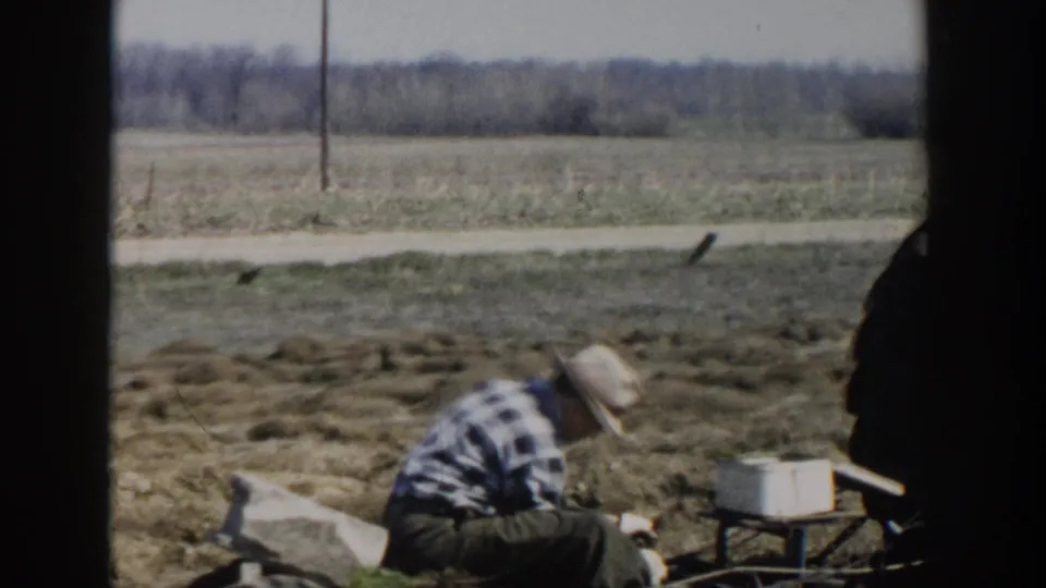 1954 Wisconsin Dells home-movie frame of a man in plaid crouching in a field, with open farmland and bare trees beyond.