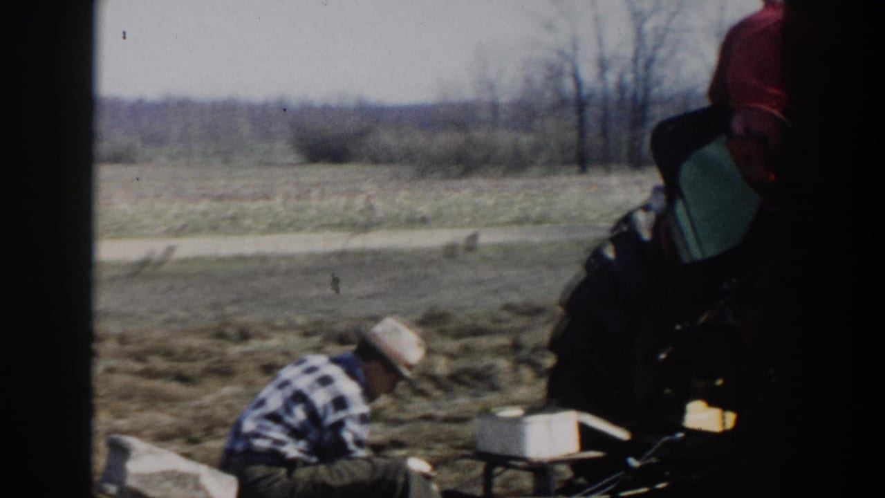 1954 Wisconsin Dells farm scene with a crouching man in a straw hat near tractor machinery and a pale rural horizon.