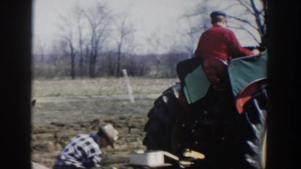 1954 Wisconsin Dells home-movie view of a red-shirted tractor operator beside a crouching field worker in tilled farmland.