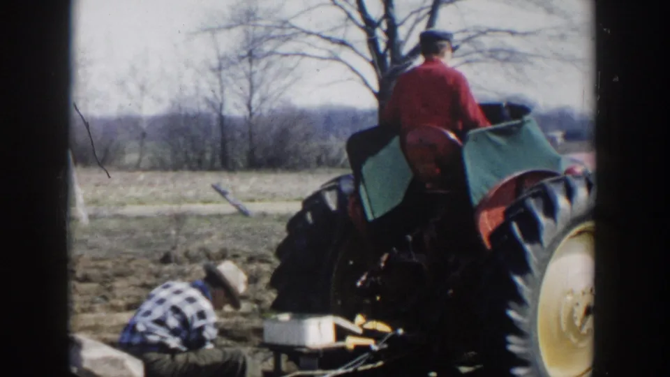 1954 Wisconsin Dells frame showing a red tractor and seated operator near a man crouched over field work, with bare trees behind.