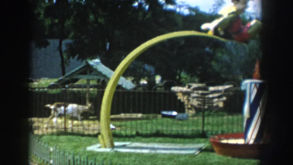 1954 Wisconsin Dells 8mm still showing a yellow arch and doll figure near a fenced deer enclosure and small shelter.