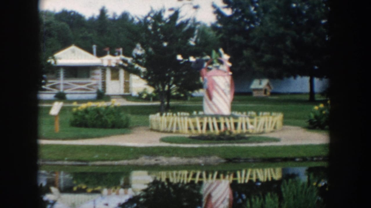 1954 Wisconsin Dells miniature park view with a candy-striped tower, fenced display, manicured grass, trees, and water in the foreground.