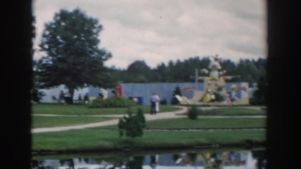 Visitors at a 1954 Wisconsin Dells miniature attraction park walk past painted displays, grass, trees, and reflective water.