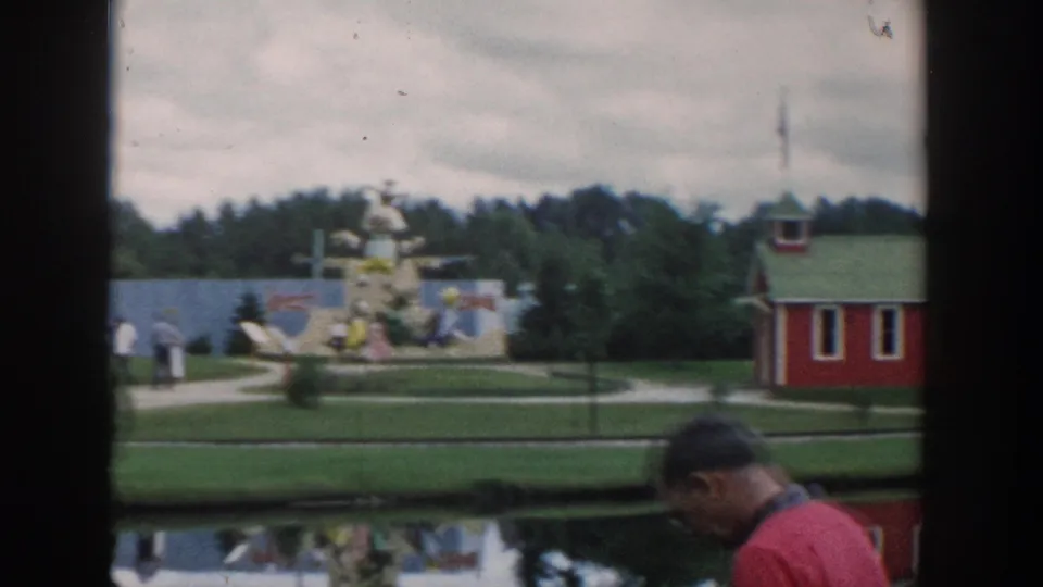 Wisconsin Dells miniature park scene with a red schoolhouse, painted displays, walkways, pond reflection, and an adult man entering the foreground.