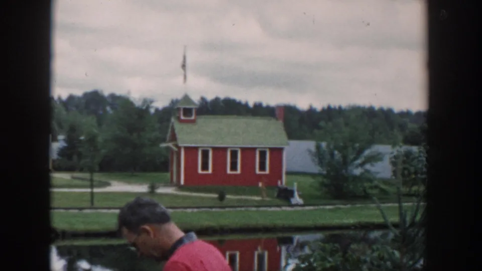 1954 Wisconsin Dells view of a red miniature schoolhouse with a green roof and flag, seen across a pond as an adult man passes in front.