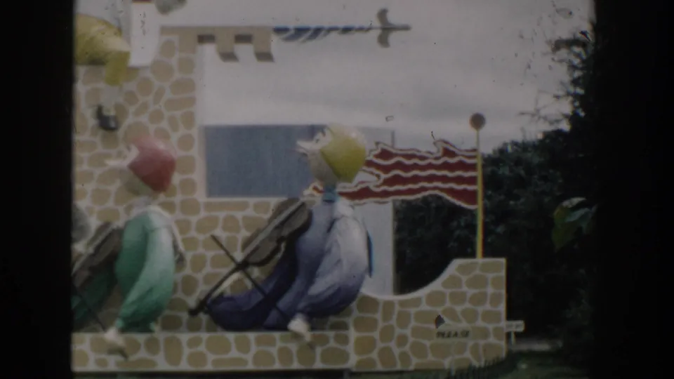 1954 Wisconsin Dells home-movie frame showing colorful violin-playing mannequins near a stone-patterned backdrop and striped flag.