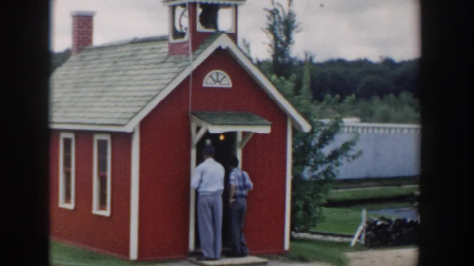 1954 Wisconsin Dells frame showing two adults at a red wooden playhouse entrance beside a lawn and waterway.