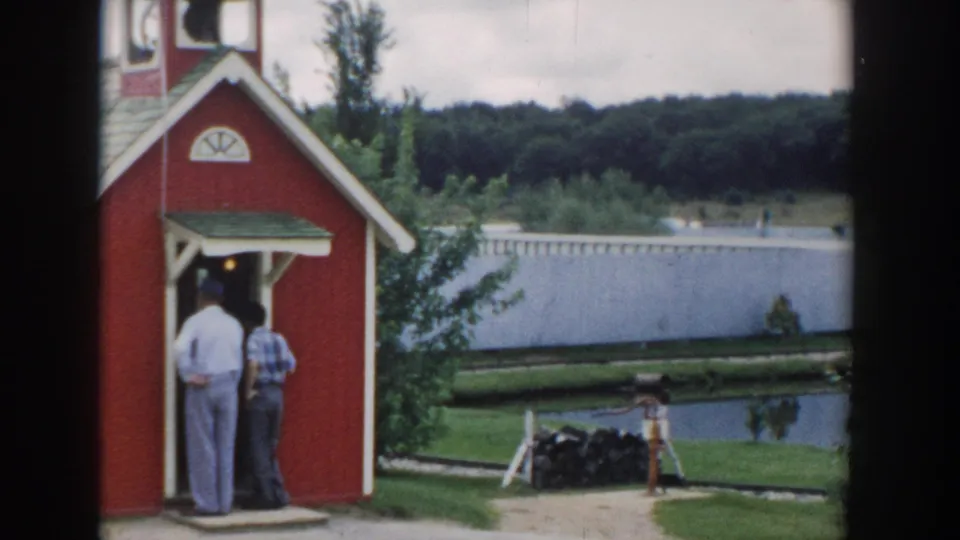 Visitor-shot 8mm frame of a red playhouse with steeple, two adults in the doorway, and a riverside backdrop in Wisconsin Dells.