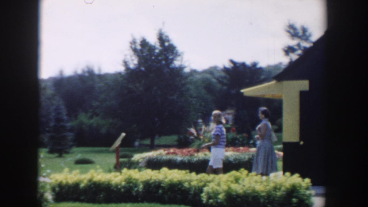 Two women in 1950s summer clothing walk through a landscaped garden in Wisconsin Dells, framed by dark window edges.