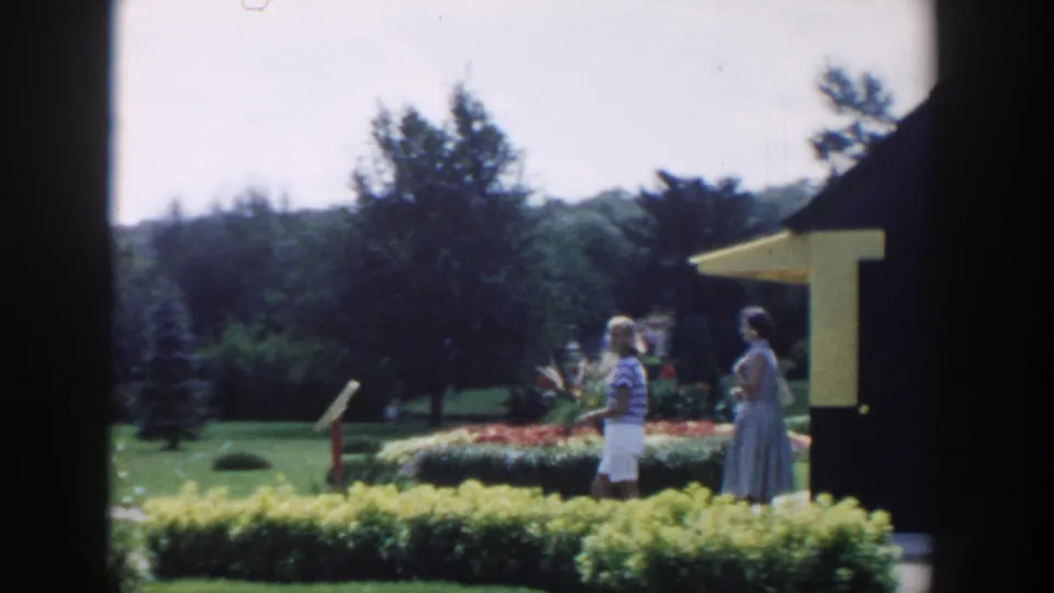 Two women in 1950s summer clothing walk through a landscaped garden in Wisconsin Dells, framed by dark window edges.