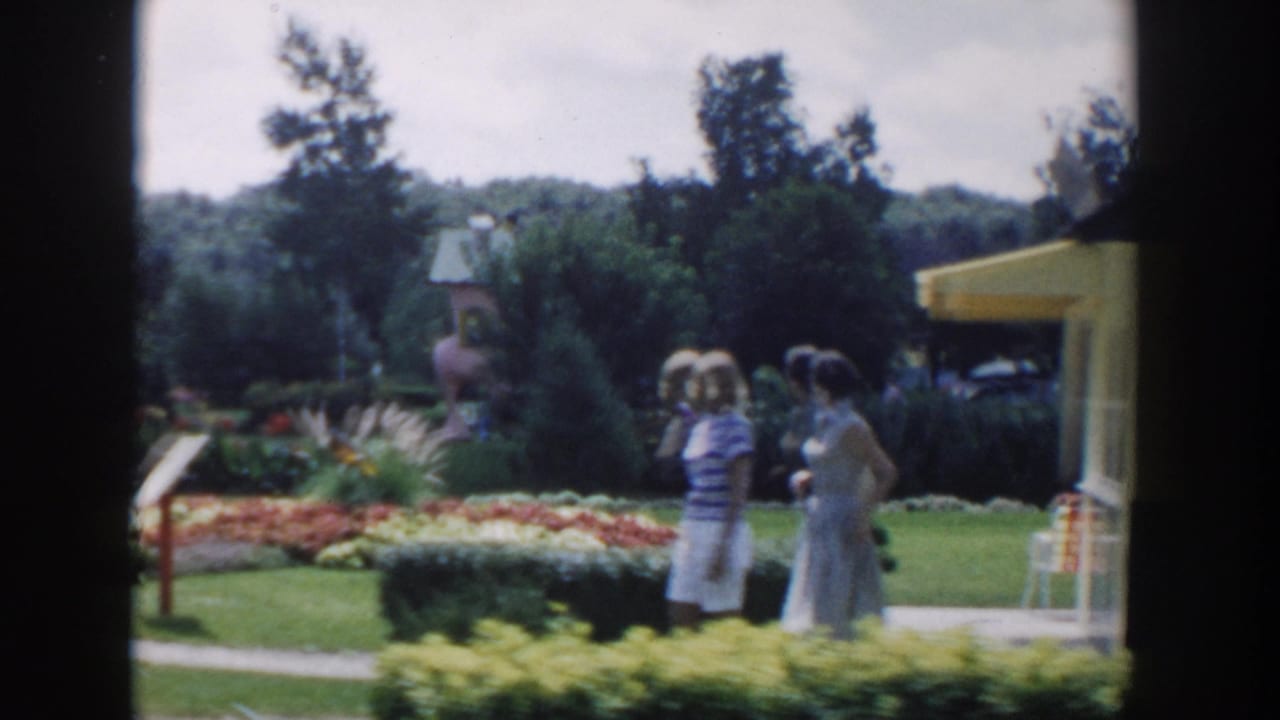 In the Wisconsin Dells garden scene, two women stand near a path bordered by flowers, hedges, lawn, and a nearby building edge.