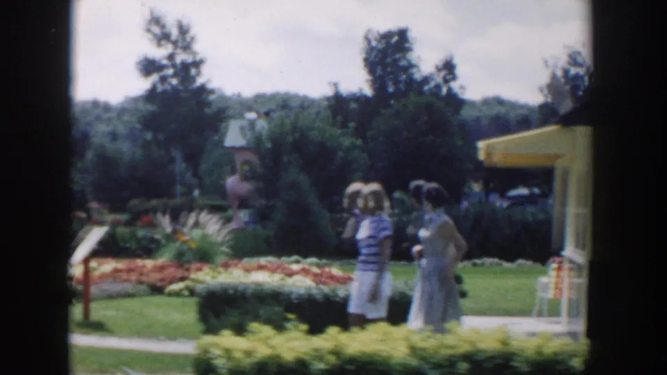 In the Wisconsin Dells garden scene, two women stand near a path bordered by flowers, hedges, lawn, and a nearby building edge.
