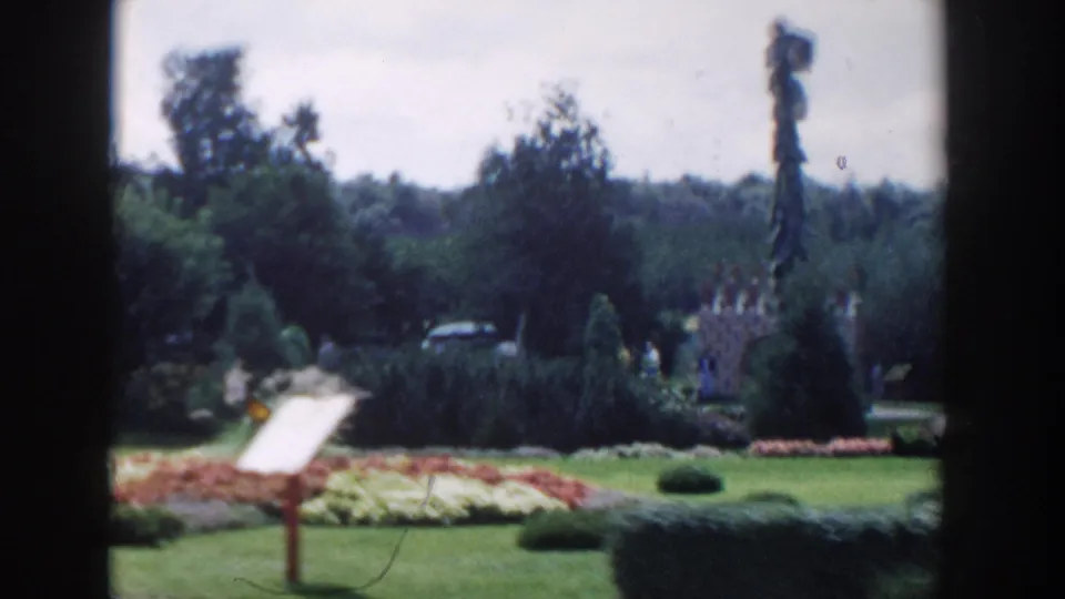 A wider Wisconsin Dells garden view shows flowerbeds, shrubs, trees, a garden sign, distant cars, and a vertical dark frame edge.