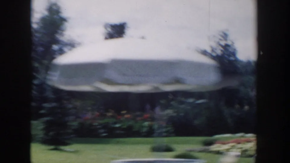A large light-colored umbrella or canopy appears in the Wisconsin Dells garden, with flowerbeds, shrubs, trees, and soft motion blur.
