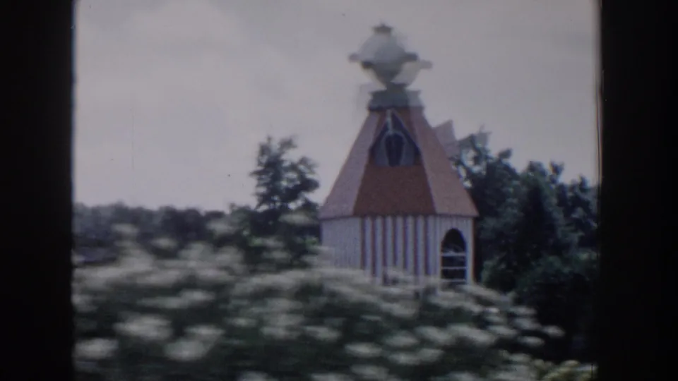 1954 Wisconsin Dells frame showing a small white striped garden building with a brown conical roof, partly screened by flowers and foliage.
