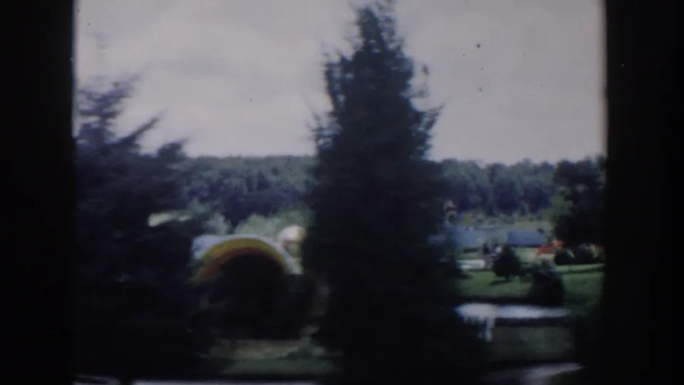 Soft home-movie frame showing a Wisconsin Dells park landscape with tall trees, lawn, flower beds, and a small curved structure or feature in the garden.