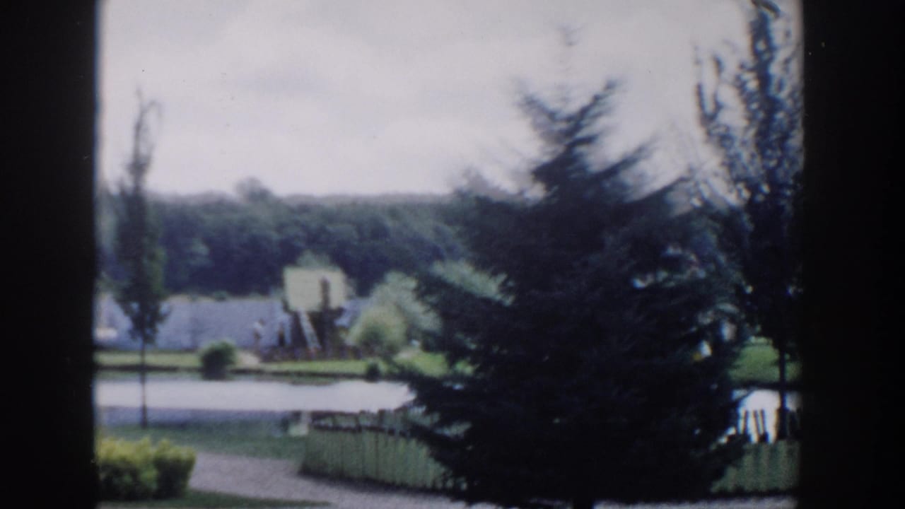 1954 Wisconsin Dells home-movie view of a landscaped lakeside park seen through dark vertical framing, with trees, lawn, water, and a small shore feature.