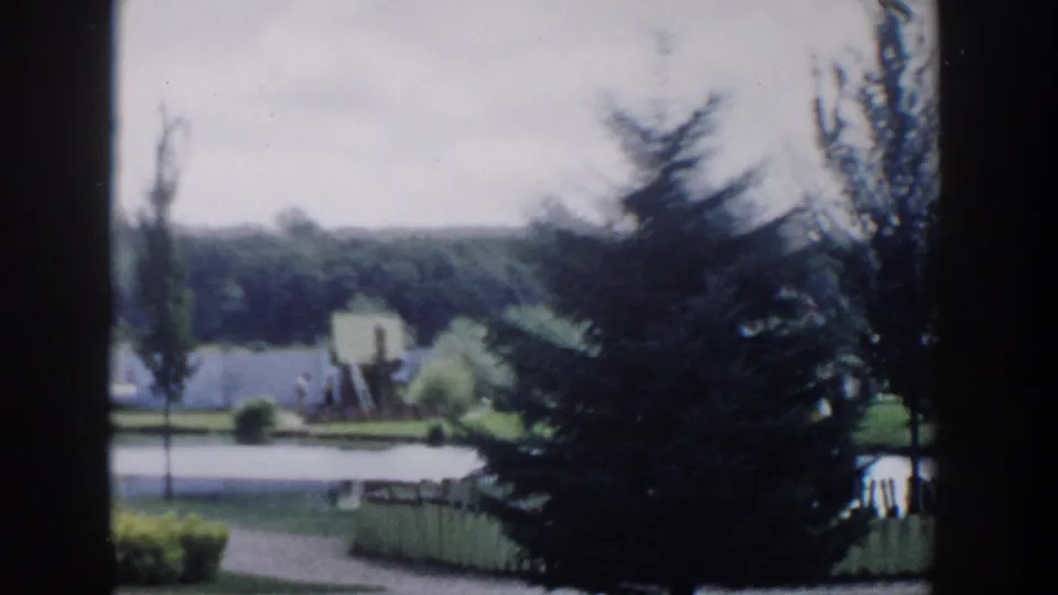 1954 Wisconsin Dells home-movie view of a landscaped lakeside park seen through dark vertical framing, with trees, lawn, water, and a small shore feature.
