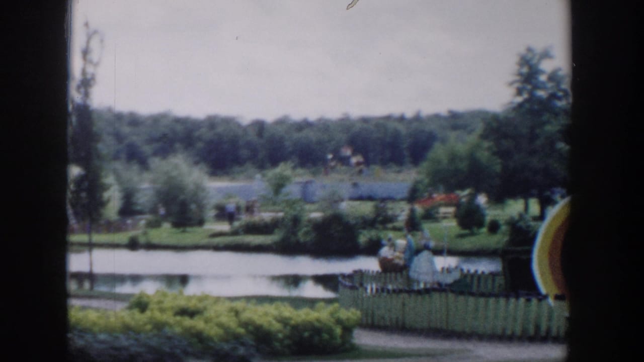 Park and lake with white fencing, distant visitors, trees, shrubs, and a large yellow curved decorative feature at the right edge.