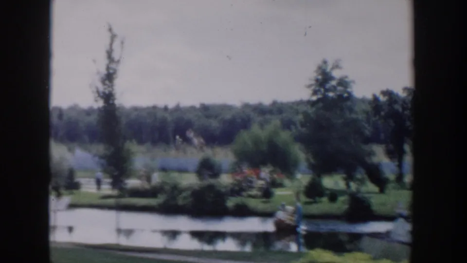 1954 Wisconsin Dells home-movie frame showing a wide landscaped park beside water, with trees, shrubs, paths, and distant visitors in soft focus.