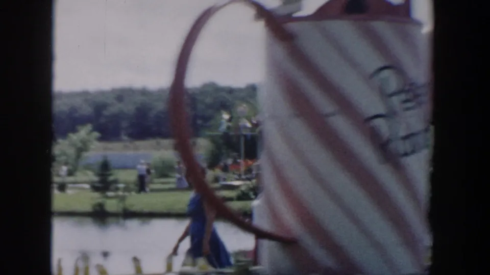 1954 Wisconsin Dells lakeside park footage with distant visitors, white fencing, lawns, trees, and a large white-and-red decorative object close to the camera.