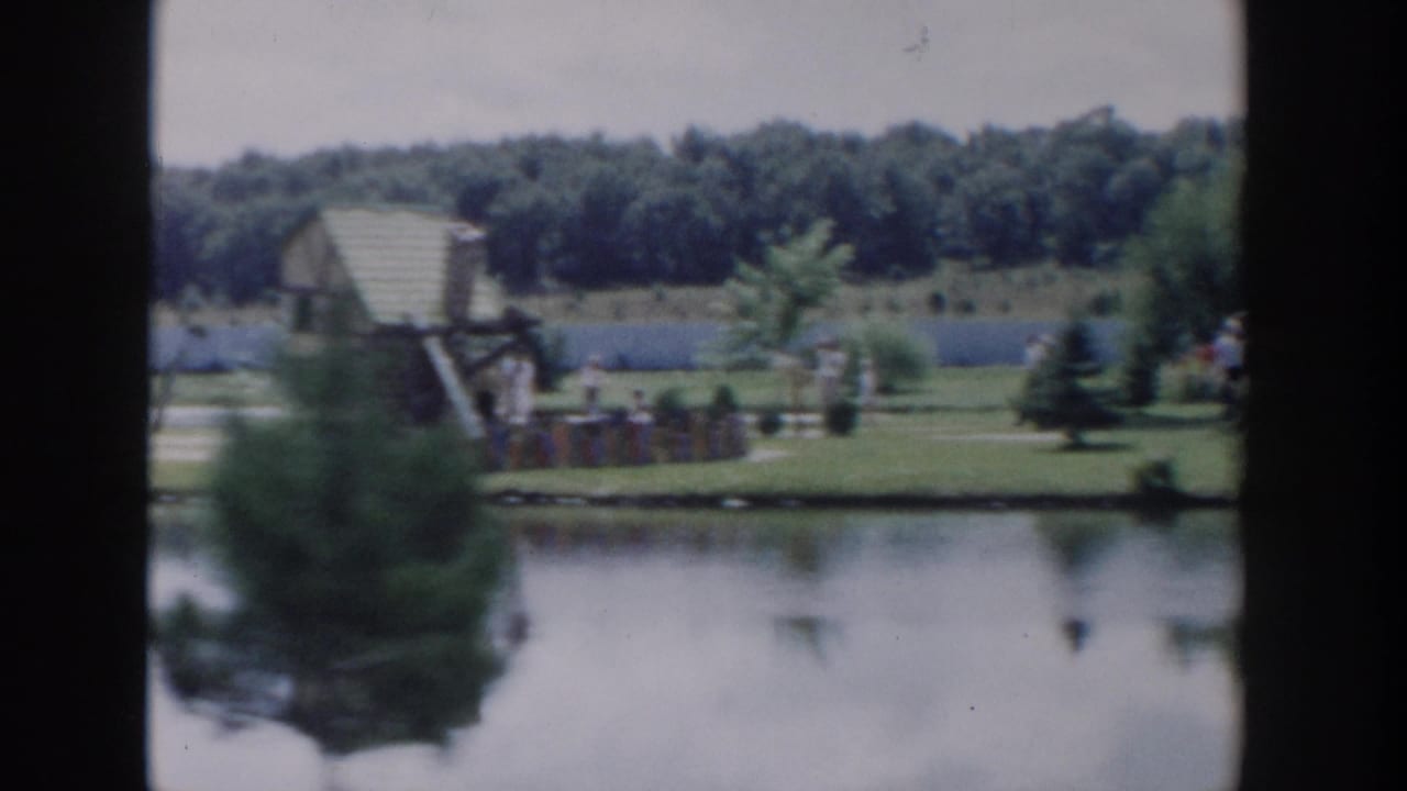 1954 Wisconsin Dells lakeside frame with a small wooden structure, people on the grass, trees, and calm water.