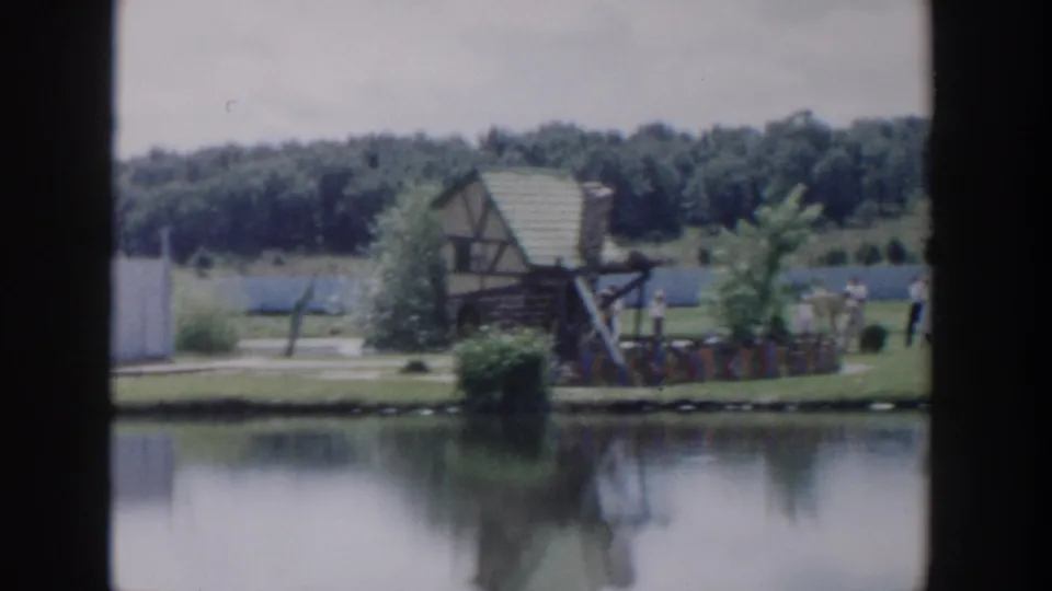 1954 Wisconsin Dells home-movie frame showing visitors near a wooden lakeside structure with shrubs and water reflections.